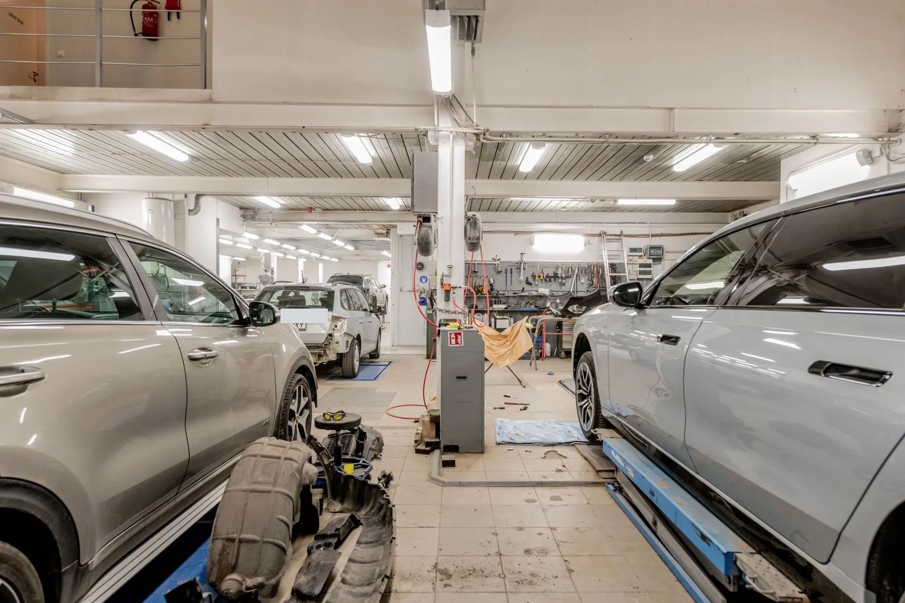 Nave de un taller con dos coches subidos en elevadores y un paragolpes desmontado en el suelo